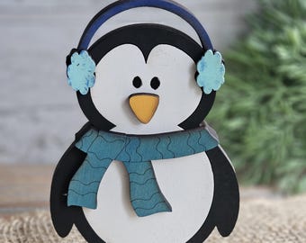 May include: A wooden penguin figurine with a black and white body, wearing blue earmuffs and a blue scarf. The penguin has a yellow beak and is standing on a blue base. The figurine is a decorative item.