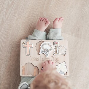 May include: A wooden puzzle with a cross, crown of thorns, chalice, and tomb. The puzzle pieces are removable and fit into the corresponding cutouts. The puzzle is personalized with the text "Jesus Loves Ziggy".