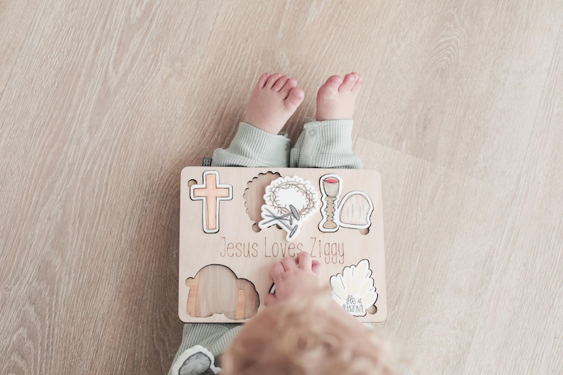 May include: A wooden puzzle with a cross, crown of thorns, chalice, and tomb. The puzzle pieces are removable and fit into the corresponding cutouts. The puzzle is personalized with the text "Jesus Loves Ziggy".
