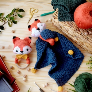 Pode incluir: Dois brinquedos de raposa de crochê, um com um anel de madeira, sobre uma superfície de madeira com um cobertor de crochê azul e novelos de lã.
