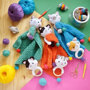 Pode incluir: Um conjunto de cinco brinquedos de gato de crochê com cobertores. Cada brinquedo de gato tem um cobertor de uma cor diferente e um chocalho. Os cobertores são verdes, laranja, azuis e brancos. Os brinquedos de gato são todos brancos com olhos e narizes pretos.