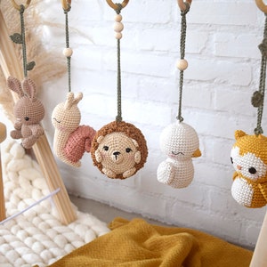 May include: A wooden baby gym with five hanging crochet toys: a bunny, snail, hedgehog, duck, and owl. The toys are in soft pastel colors, suspended from wooden rings with green cords. The background includes a white brick wall and a yellow blanket.