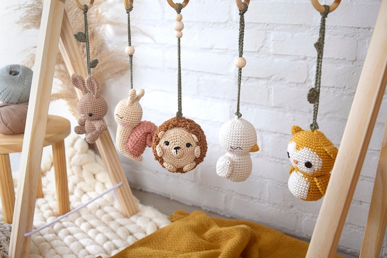 May include: A wooden baby gym with five hanging crochet toys: a bunny, snail, hedgehog, duck, and owl. The toys are in soft pastel colors, suspended from wooden rings with green cords. The background includes a white brick wall and a yellow blanket.