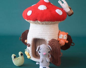 May include: A crocheted mushroom house with a red and white top, a white stem, and a brown door. There are four crocheted animals near the house: a frog, a mouse, a squirrel, and a bunny.