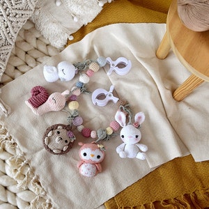 May include: A handmade crochet baby mobile featuring a white bunny, owl, snail, hedgehog, and a swan. The mobile is adorned with pastel-colored beads and white plastic clips. The items are arranged on a beige blanket.