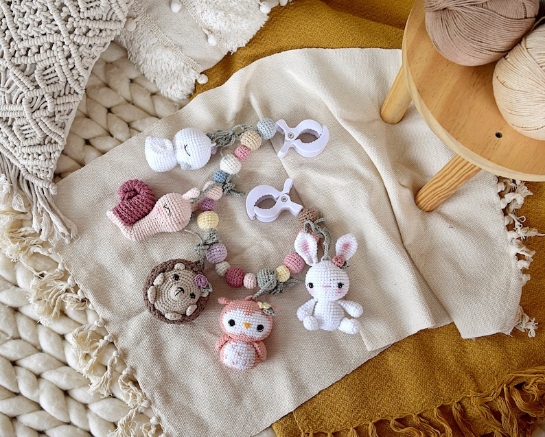 May include: A handmade crochet baby mobile featuring a white bunny, owl, snail, hedgehog, and a swan. The mobile is adorned with pastel-colored beads and white plastic clips. The items are arranged on a beige blanket.