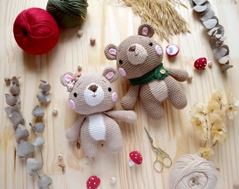 May include: Two crocheted teddy bears, one with a green scarf and the other with a pink flower crown, on a wooden surface with yarn, beads, and dried flowers.