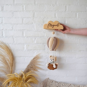 May include: A wooden cloud-shaped sign with the name "Miguel" engraved on it hangs from a white brick wall. A crocheted hot air balloon with brown and beige stripes is attached to the sign and carries a crocheted teddy bear.