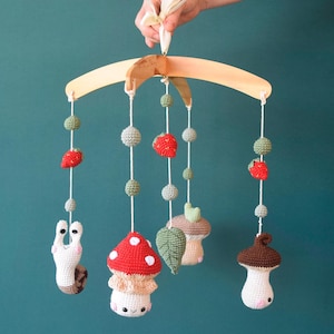 May include: A wooden baby mobile with crocheted hanging toys. The toys include a snail, two mushrooms, a leaf, and two strawberries. The mobile is suspended from a wooden arch.
