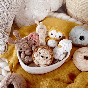 May include: A collection of handmade crochet animal toys, including a bunny, snail, owl, lion, and bear, nestled in a white woven bowl on a mustard-yellow fabric. Several balls of yarn in neutral colors are also visible.