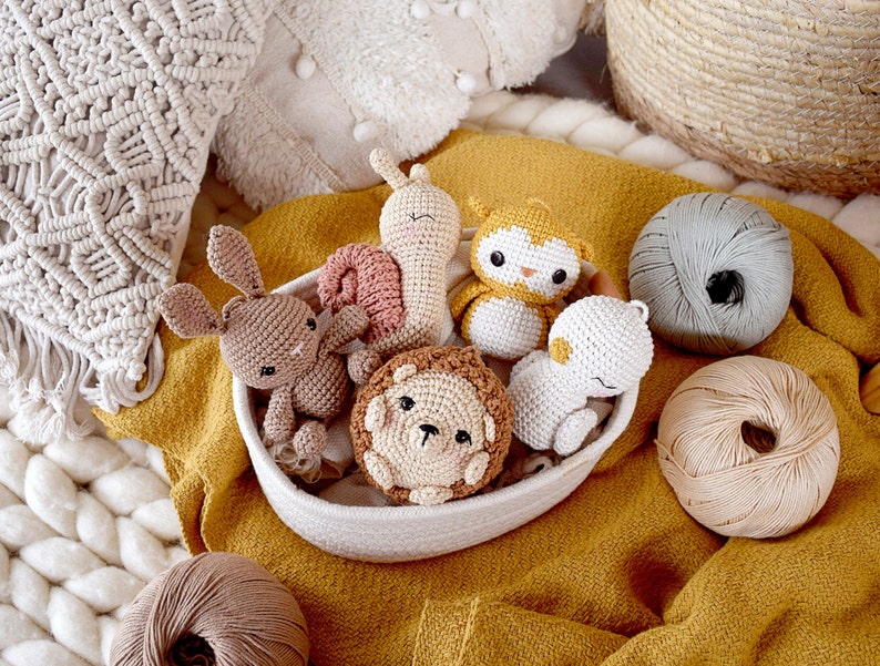 May include: A collection of handmade crochet animal toys, including a bunny, snail, owl, lion, and bear, nestled in a white woven bowl on a mustard-yellow fabric. Several balls of yarn in neutral colors are also visible.