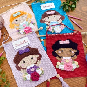 Pode incluir: Três tapeçarias de parede de crochê com uma menina com flores. As tapeçarias são rosa, azul e vermelho. Os nomes "Isabella", "Catalina" e um terceiro nome estão escritos nas tapeçarias.