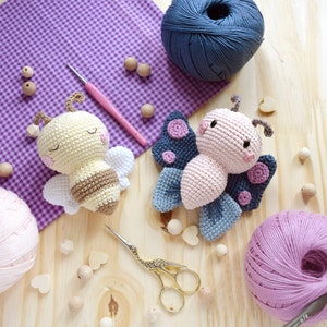 Pode incluir: Um close-up de dois brinquedos amigurumi de crochê, uma abelha e uma borboleta, sobre uma superfície de madeira com novelos de lã e contas de madeira. A abelha é amarela e marrom com listras pretas, e a borboleta é rosa com asas azuis e roxas.