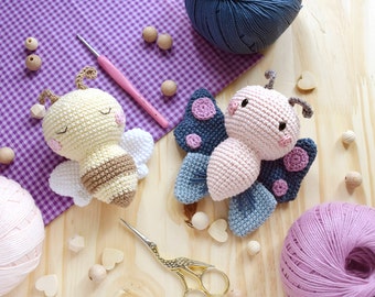 May include: A close-up of two crocheted amigurumi toys, a bee and a butterfly, on a wooden surface with yarn balls and wooden beads. The bee is yellow and brown with black stripes, and the butterfly is pink with blue and purple wings.