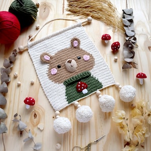Pode incluir: Uma tapeçaria de parede de crochê feita à mão com um urso marrom com bochechas rosadas e um cachecol verde. O urso está em um fundo branco, com contas de madeira e pompons brancos. Pequenos detalhes de cogumelos vermelhos e brancos também estão incluídos.
