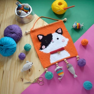 Pode incluir: Um pendurado de parede de crochê com um gato com rosto preto e branco, fundo laranja e fio branco para o corpo do gato. O gato tem uma coleira vermelha com um sino. O pendurado tem uma variedade de peixes de crochê e outros pequenos brinquedos de crochê pendurados nele.