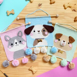 Pode incluir: Três tapeçarias de parede de crochê com diferentes raças de cães. Cada tapeçaria é feita com um fundo azul claro, rosa ou verde e apresenta um cão com pelo castanho, cinza ou bege. Cada tapeçaria tem um amuleto dourado e é decorada com pompons fofinhos.