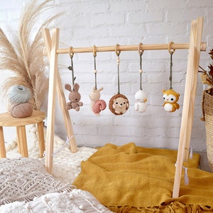 May include: A wooden baby gym with five hanging crochet toys: a bunny, a snail, a hedgehog, a bird, and an owl. The toys are in neutral colors, with wooden beads and rings. The gym is set on a yellow blanket.