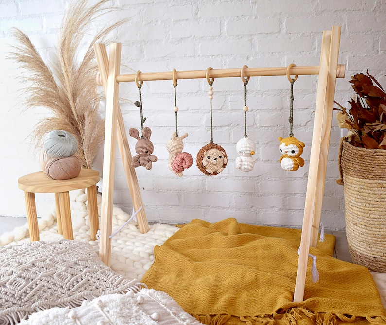 May include: A wooden baby gym with five hanging crochet toys: a bunny, a snail, a hedgehog, a bird, and an owl. The toys are in neutral colors, with wooden beads and rings. The gym is set on a yellow blanket.