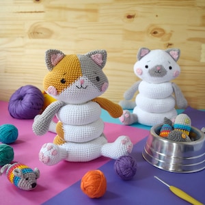 Op de afbeelding: Twee gehaakte katten speelgoedjes, een oranje, wit en grijs, en een witte, zitten op een roze en paarse ondergrond. De oranje kat zit rechtop met zijn poten uitgestrekt. De witte kat zit met zijn poten onder zich. Er zijn verschillende bollen garen en een gehaakt vis speelgoedje op de achtergrond.