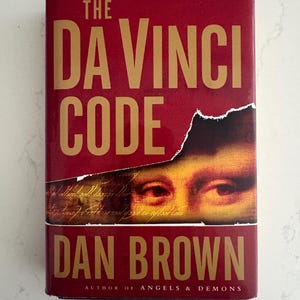 May include: A burgundy hardcover book, "The Da Vinci Code" by Dan Brown, with gold lettering. The cover features a torn section revealing an image of eyes. The author is also the author of "Angels & Demons".