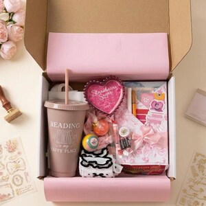 Puede incluir: Una caja de regalo llena de art&iacute;culos rosas y blancos, que incluyen un vaso con el texto "Reading is my happy place", un recipiente en forma de coraz&oacute;n con el texto "Strawberry Sugar", pegatinas y un pastel peque&ntilde;o. Tambi&eacute;n se ve un ramo de flores rosas y un sello de madera.