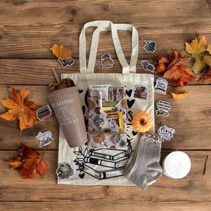 May include: A cream-coloured canvas tote bag featuring a black line drawing of stacked books, a mauve tumbler with the text Reading is my happy place, a patterned fabric pouch, a pair of grey socks, a small orange pumpkin, and various book-themed stickers.