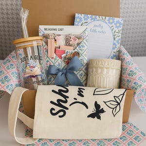Pode incluir: Uma caixa de presente contendo um copo de vidro com tampa de bambu e canudo, impresso com o texto 'JUST A GIRL WHO LOVES BOOKS', um envelope 'READING LIST', um bloco de notas 'pretty little notes', uma vela creme texturizada e uma sacola.