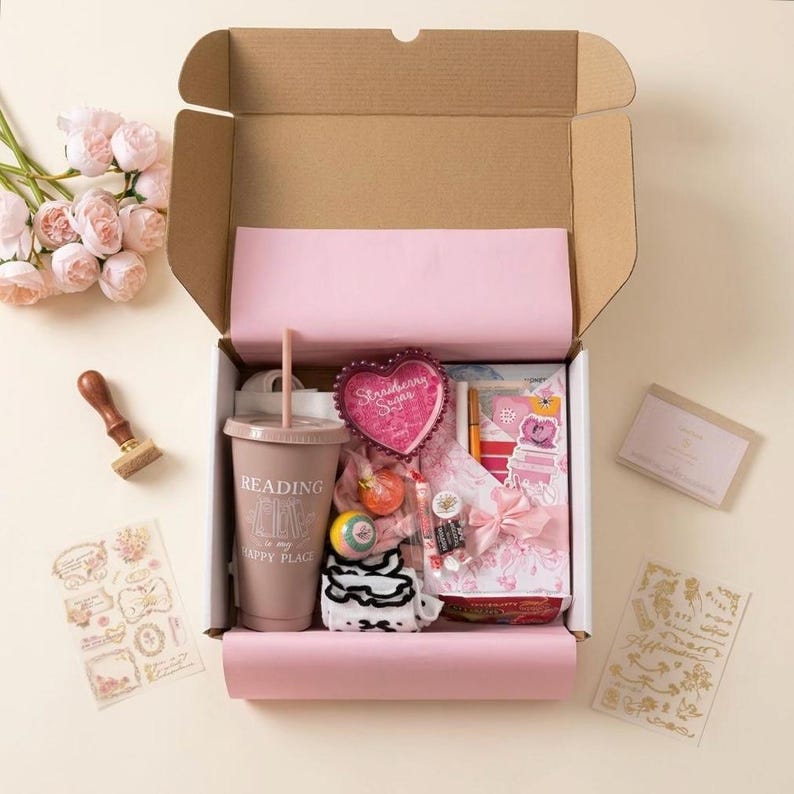 Puede incluir: Una caja de regalo llena de art&iacute;culos rosas y blancos, que incluyen un vaso con el texto "Reading is my happy place", un recipiente en forma de coraz&oacute;n con el texto "Strawberry Sugar", pegatinas y un pastel peque&ntilde;o. Tambi&eacute;n se ve un ramo de flores rosas y un sello de madera.