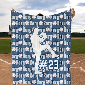 May include: A blue and white baseball themed blanket with the name "Rory" repeated throughout the design. A white silhouette of a baseball player is in the center of the blanket with the number "#23" on the front of the jersey.