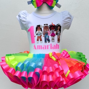 May include: A white t-shirt with a graphic of four cartoon girls and the number 1. The shirt says "Amaria" in pink glitter. The shirt is paired with a rainbow tulle tutu with pink, blue, green, and yellow ribbon.