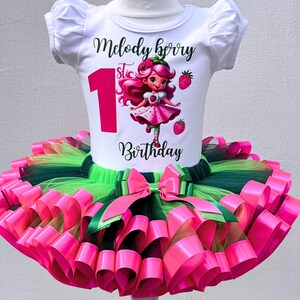 May include: A white short-sleeved shirt with the text "Melody Berry 1st Birthday" and a cartoon strawberry girl design. The shirt is paired with a layered tutu skirt in pink, green, and black, with a large pink bow.