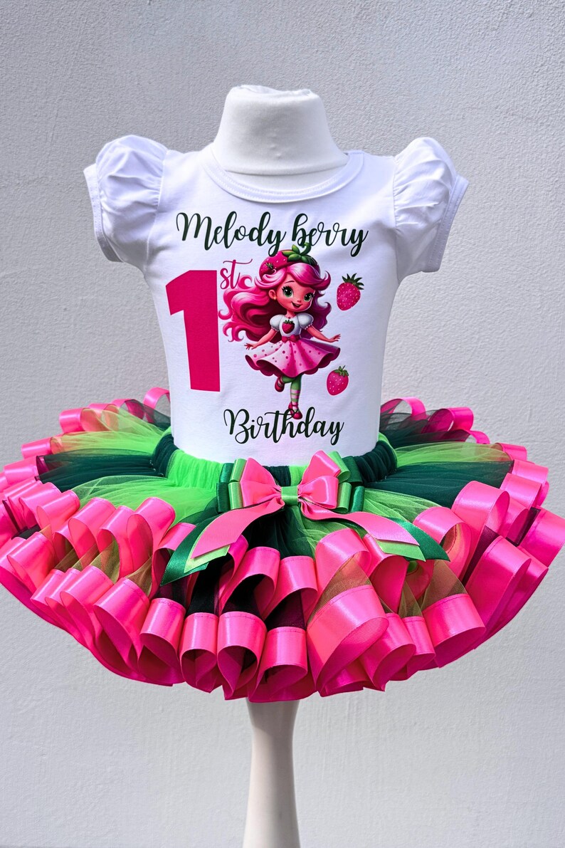 May include: A white short-sleeved shirt with the text "Melody Berry 1st Birthday" and a cartoon strawberry girl design. The shirt is paired with a layered tutu skirt in pink, green, and black, with a large pink bow.