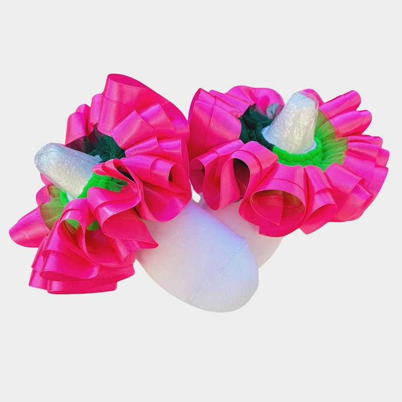 May include: Two white objects are adorned with vibrant fuchsia pink ribbon, green accents, and a white cone-shaped element. The ribbon is arranged in a ruffled, layered design, creating a bold and eye-catching aesthetic.