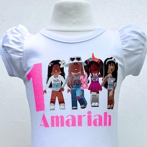 May include: White t-shirt with a pink number one and the name "Amaria" printed on it. The shirt features a graphic of four cartoon girls with different hairstyles and outfits.