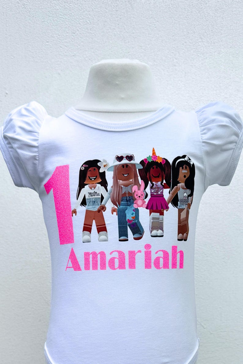 May include: White t-shirt with a pink number one and the name "Amaria" printed on it. The shirt features a graphic of four cartoon girls with different hairstyles and outfits.