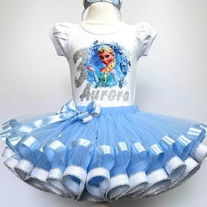 May include: A white t-shirt with a picture of Elsa from Frozen, the number 3, and the name "Aurora" in silver glitter. The shirt is paired with a light blue tulle tutu with light blue satin ribbon trim and a silver crown.