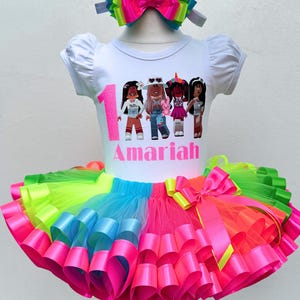 May include: A white t-shirt with a colorful graphic of cartoon girls and the number 1. The shirt says "Amaria" in pink letters. The shirt is paired with a rainbow tulle tutu with pink, blue, green, and yellow ribbons.