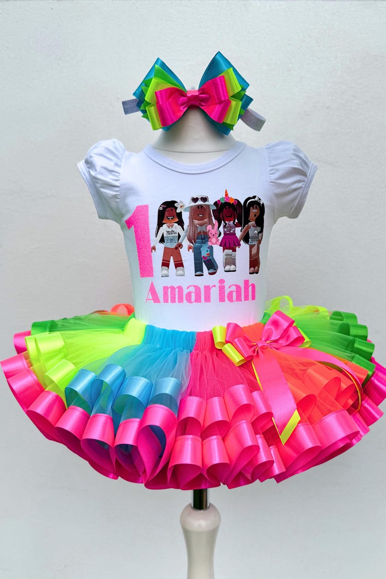 May include: A white t-shirt with a colorful graphic of cartoon girls and the number 1. The shirt says "Amaria" in pink letters. The shirt is paired with a rainbow tulle tutu with pink, blue, green, and yellow ribbons.
