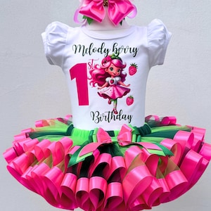 May include: A white t-shirt with the text "Melody Berry 1st Birthday" and a cartoon strawberry girl design. The outfit includes a pink and green tutu skirt and a matching bow headband. The outfit is designed for a first birthday celebration.