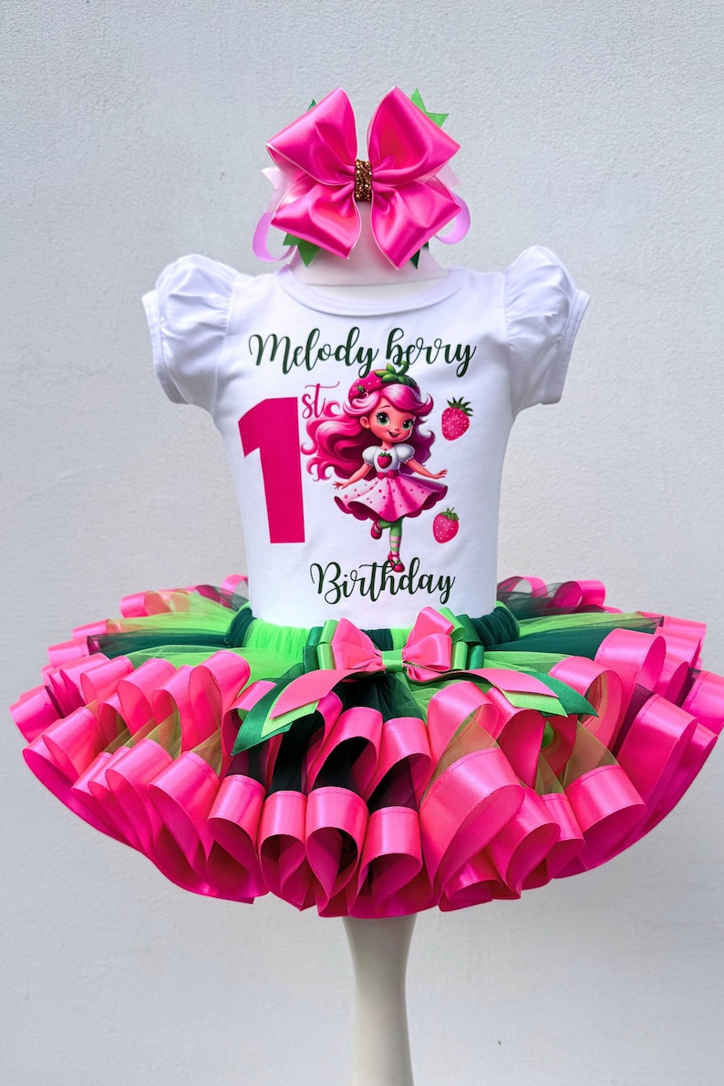 May include: A white t-shirt with the text "Melody Berry 1st Birthday" and a cartoon strawberry girl design. The outfit includes a pink and green tutu skirt and a matching bow headband. The outfit is designed for a first birthday celebration.