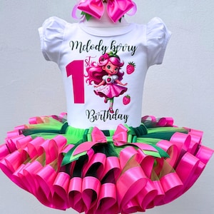 May include: A white t-shirt with a graphic of a cartoon girl and the text "Melody Berry 1st Birthday" paired with a pink tutu skirt and matching bow. The tutu is layered with pink and green ribbons. A festive outfit for a first birthday.