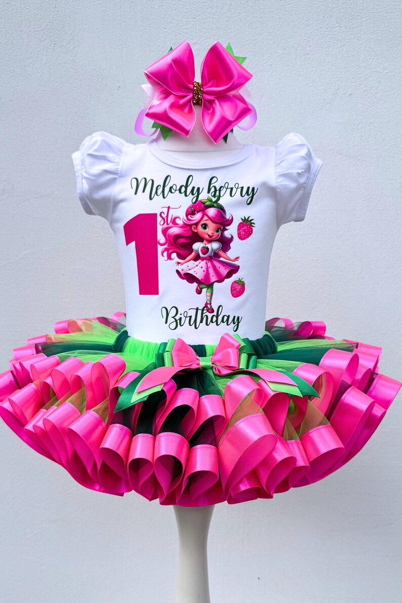 May include: A white t-shirt with a graphic of a cartoon girl and the text "Melody Berry 1st Birthday" paired with a pink tutu skirt and matching bow. The tutu is layered with pink and green ribbons. A festive outfit for a first birthday.