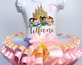 May include: A white t-shirt with a gold glitter castle and Disney Princesses, the number 1, and the name "tiffani" in gold glitter. The shirt is paired with a rainbow tulle tutu with pink satin ribbon trim.