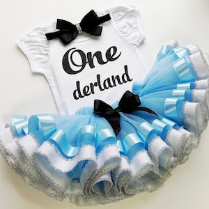 May include: A light blue tulle tutu skirt with white and blue ribbon trim. A white onesie with black glitter lettering that says "One derland" is included. A black bow headband is also included.