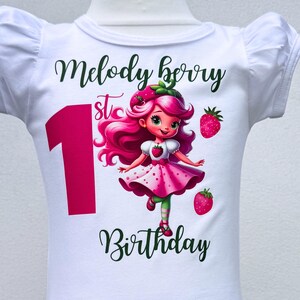 May include: White baby bodysuit with a graphic of a strawberry girl and the text "Melody Berry 1st Birthday". The number 1 is in pink, and the text is in green. The strawberry girl has pink hair and a pink dress.
