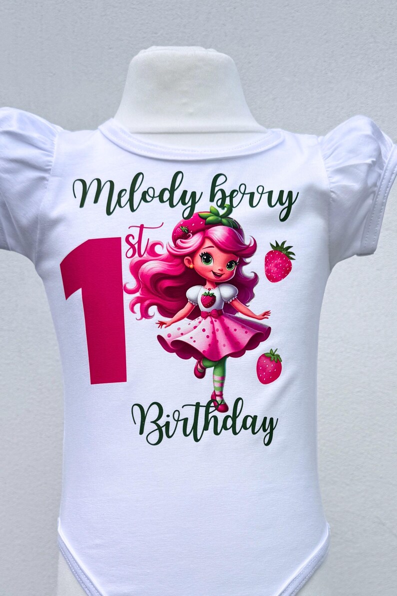 May include: White baby bodysuit with a graphic of a strawberry girl and the text "Melody Berry 1st Birthday". The number 1 is in pink, and the text is in green. The strawberry girl has pink hair and a pink dress.