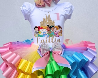 以下が含まれることがあります： 金色の城とプリンセスのグラフィック、数字の1、そして「Caitlin」という名前が金色のグリッターで描かれた白い半袖トップス。虹色のチュチュは、カラフルなリボンの層とラインストーンの大きなリボンが付いています。お揃いのリボンヘアバンドがこの衣装を完成させます。