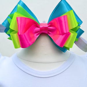May include: A large, multi-colored bow made of satin ribbon. The bow is layered with turquoise, lime green, and hot pink ribbon. The bow is tied with a white knot in the center.