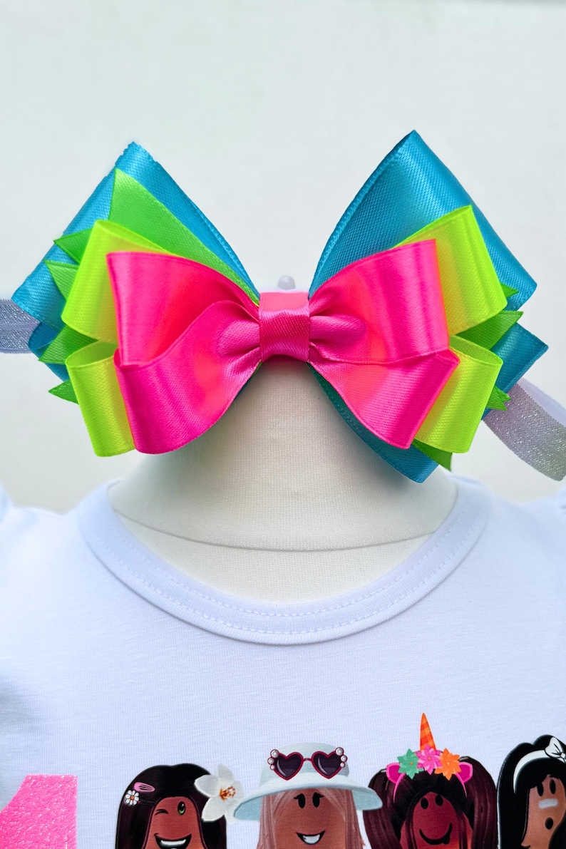 May include: A large, multi-colored bow made of satin ribbon. The bow is layered with turquoise, lime green, and hot pink ribbon. The bow is tied with a white knot in the center.